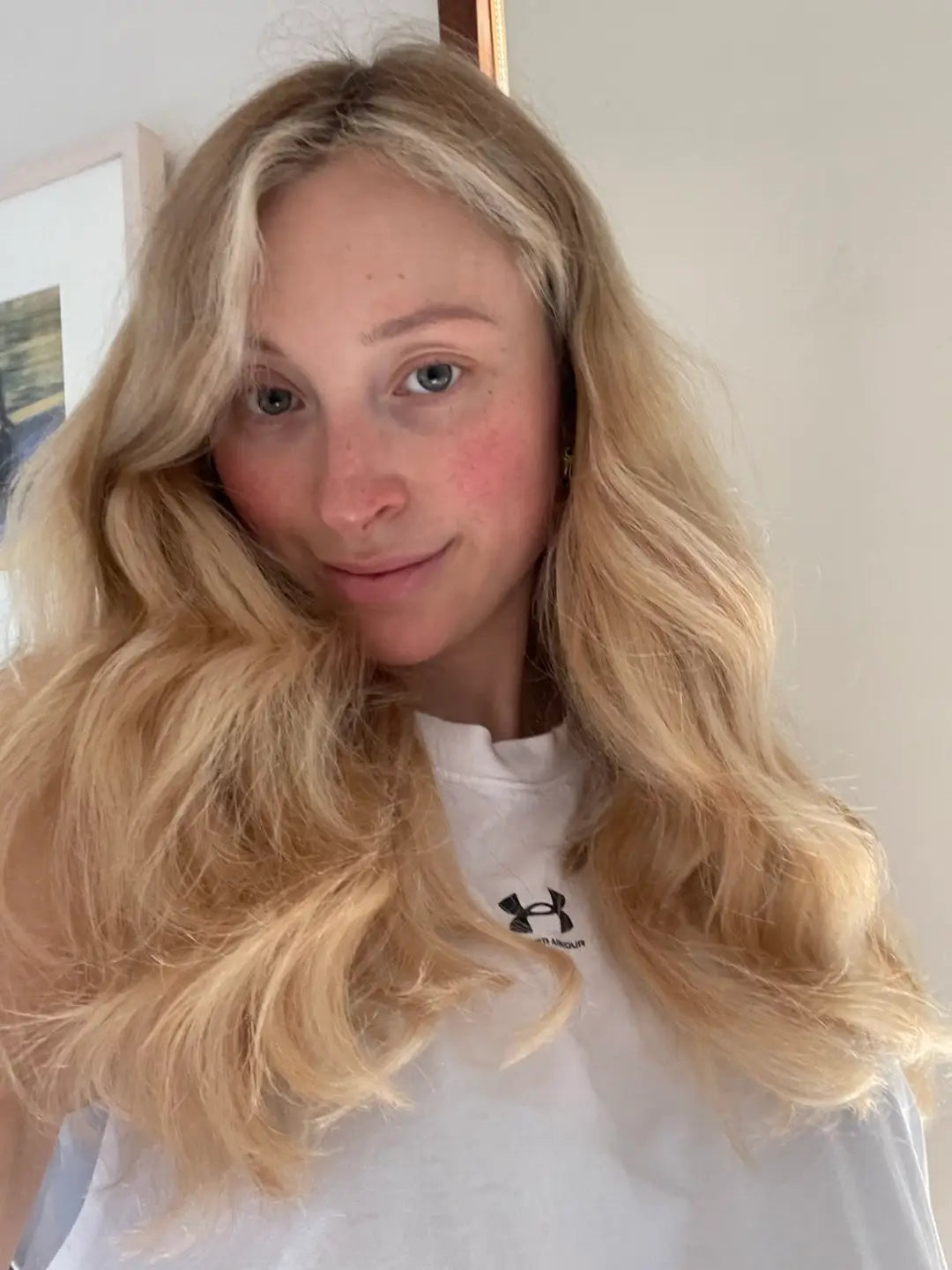 Woman with long, voluminous wavy blonde hair wearing a white shirt, smiling softly in natural indoor light.